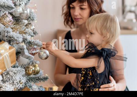 Eine Mutter mit einer 2-jährigen Tochter schmückt den Weihnachtsbaum. Beide tragen schwarze Kleider, die Tochter hängt einen Ball an den Weihnachtsbaum. Stockfoto