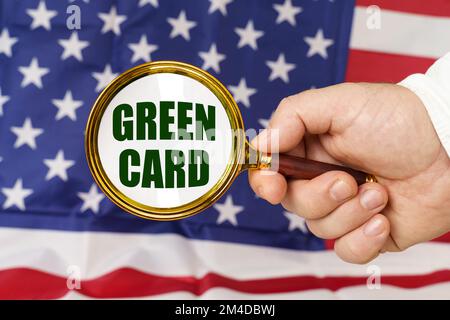 Vor der amerikanischen Flagge hält ein Mann eine Lupe in der Hand mit der Inschrift Green Card. Stockfoto