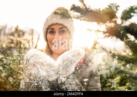 Glückliche Frau in Winterkleidung, die an einem verschneiten Wintertag im Freien Schnee bläst. Stockfoto