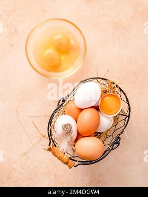 Verschiedene Eier im Metallkorb auf beigefarbenem Hintergrund. Draufsicht auf rohe braune Eier und weiße Eier. Rohe zerbrochene Eier in einer Glasschüssel. Stockfoto