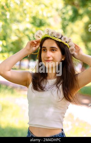 Glückliches Mädchen mit Kranz aus weißem Elendelion auf grünem Rasen am sonnigen Sommertag. Hochwertige Fotos Stockfoto