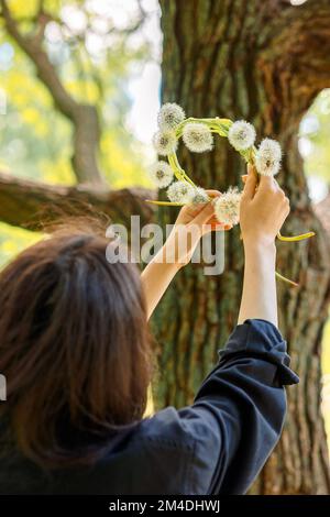 Glückliches Mädchen mit Kranz aus weißem Elendelion auf grünem Rasen am sonnigen Sommertag. Hochwertige Fotos Stockfoto