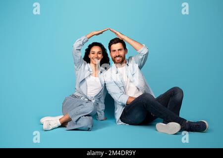Fröhliches junges europäisches Paar in zwangloser Atmosphäre machen Dachschild mit Händen, sitzen auf dem Boden, isoliert auf blauem Hintergrund Stockfoto