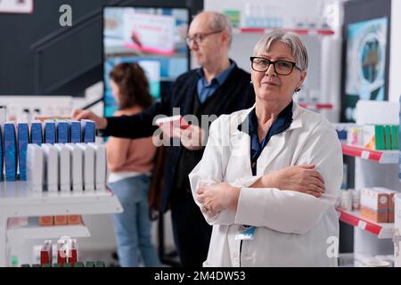 Porträt eines Chefapothekers, der in der Apotheke mit gekreuzten Armen vor die Kamera schaut und Kunden unterstützt. Kunden, die in Apothekenregalen Medikamente und Vitamine kaufen Stockfoto