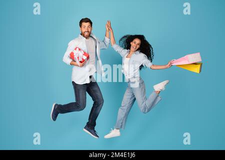 Fröhlich aufgeregt überraschte junge europäische männliche und weibliche in lässiger Form mit Geschenk, Taschen springen, in der Luft gefrieren, High Five geben Stockfoto