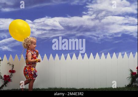 Kleines Mädchen mit lockigem Haar, trägt ein Sommerkostüm, schlendert entlang der Vorderseite eines weißen Lattenzauns und trägt einen gelben Ballon Stockfoto