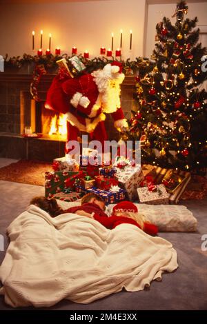 Der Weihnachtsmann steht vor dem Kamin mit Paketen auf dem Rücken und stellt sie unter den Baum, während drei Kinder auf dem Boden schlafen Stockfoto