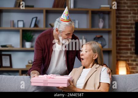 Fürsorglicher alter Ehemann, der aufgeregt reife Frau ein romantisches Geburtstagsgeschenk gibt, liebevoller älterer Mann, eine glückliche Frau mittleren Alters, eine rosa Schachtel, einen Kuss Stockfoto