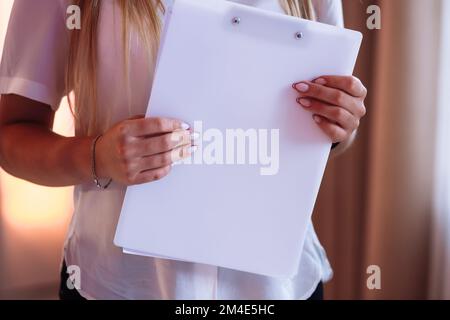 Zugeschnittenes Foto einer jungen Frau, die eine Zeitschrift oder einen Ordner mit Modell in der Hand hat. Ein Mädchen in einem weißen Hemd, das drinnen am Fenster steht Stockfoto