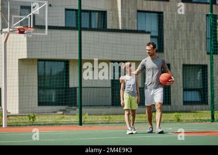 Ein langes Porträt von glücklichem Vater und Sohn, die zusammen Basketball spielen und im Sonnenlicht auf dem Platz stehen, Kopierraum Stockfoto