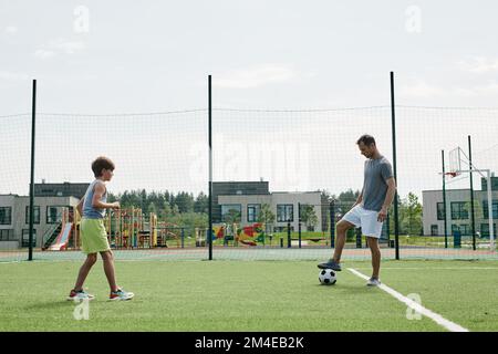 Seitenansicht von Vater und Sohn, die zusammen auf dem Freiluftplatz des modernen Wohnkomplexes Fußball spielen Stockfoto