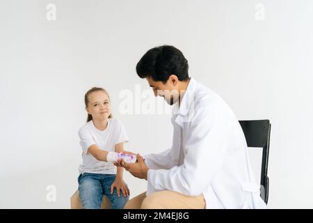 Studioaufnahme eines lächelnden verletzten kleinen Mädchens mit gebrochener Hand, eingewickelt in weißen Pflasterverband, medizinische Versorgung von freundlichem männlichen Arzt. Stockfoto