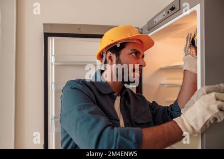 Männlicher Techniker in Uniform mit Schraubenzieher, der Kühlschrank in der Küche repariert Stockfoto