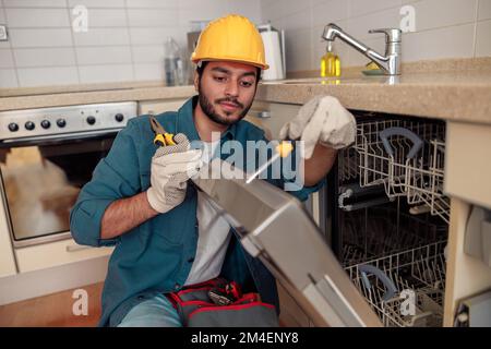 Fokussierter Handwerker in Spezialkleidung, der Geschirrspüler in der modernen Küche repariert Stockfoto