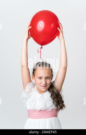 Das verspielte Mädchen im weißen Kleid hält den roten Ballon mit beiden Händen über dem Kopf und sieht aus wie eine Kamera. Studiofoto eines kaukasischen Mädchens, das zehn Jahre alt ist Stockfoto