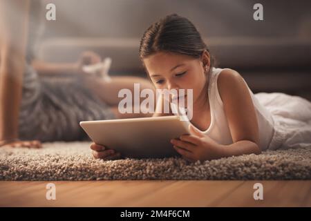 Eine App pro Tag hält Langeweile fern. Ein bezauberndes kleines Mädchen, das zu Hause ein digitales Tablet auf dem Boden mit seiner Mutter im Hintergrund benutzt. Stockfoto