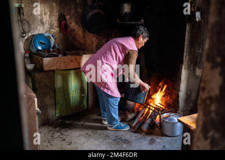 Eine Frau, die am offenen Feuer in einer kleinen, altmodischen Küche in einem Landhaus kocht Stockfoto