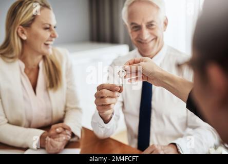Hier sind deine Schlüssel zum Schloss. Ein fröhliches, reifes Paar, das Schlüssel von einem Immobilienmakler zu seinem neuen Haus erhält, während es an einem Tisch sitzt Stockfoto