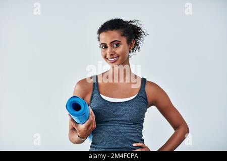 Ich bin glücklich, gesund und zu allem bereit. Eine attraktive junge Frau, die mit ihrer Yogamatte vor grauem Hintergrund posiert. Stockfoto