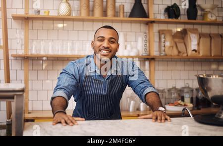 Hier ist immer Kaffeezeit. Ein gutaussehender junger Geschäftsmann, der hinter der Theke in seinem Café steht und lächelt. Stockfoto