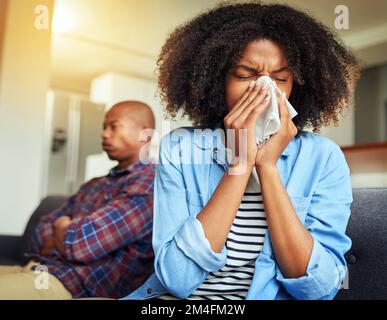 Ich hasse es, an solchen Tagen krank zu sein. Eine unbehagliche junge Frau, die in ein Taschentuch niest, während sie zu Hause auf einer Couch sitzt. Stockfoto