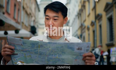 Männlicher asiatischer Tourist, der in einer neuen Stadt unterwegs ist und draußen steht, auf der Suche nach dem Ort eine Karte in Papierform anschaut, mit einem Reiseführer, um Wegbeschreibungen zu erhalten junger Mann Stockfoto