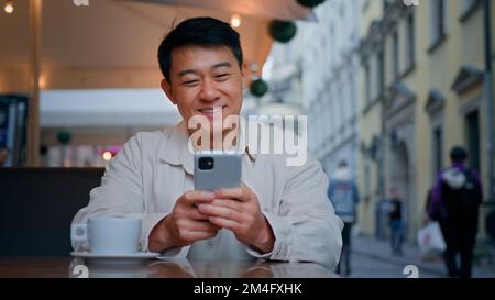 Ein lächelnder asiatischer Tourist, der im Straßencafé sitzt und Handytickets online bucht, kommuniziert per Fernzugriff mit Freunden in sozialen Netzwerken auf Stockfoto