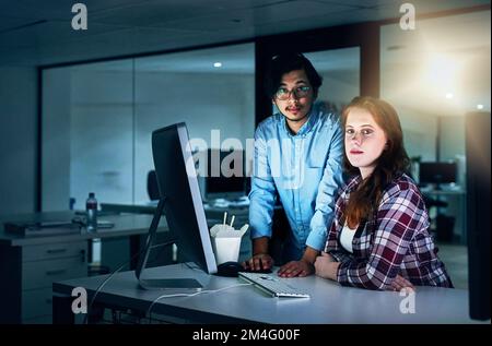 Sie arbeiten spät, damit wir besser schlafen können. Porträt von zwei fokussierten jungen Programmierern, die während der Arbeit an einem Computer im Büro zusammenarbeiten Stockfoto