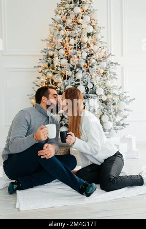 Ein verliebtes Paar umarmt und küsst sich in der Nähe des Weihnachtsbaums im dekorierten Inneren des Weihnachtsbaums zu Hause an einem Weihnachtsurlaub im Winter. Ein Mann Stockfoto