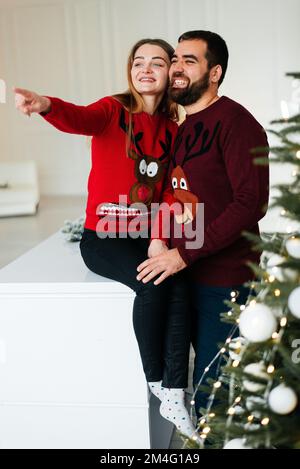 Ein verliebtes Paar umarmt und küsst sich in der Nähe des Weihnachtsbaums im dekorierten Inneren des Weihnachtsbaums zu Hause an einem Weihnachtsurlaub im Winter. Ein Mann Stockfoto