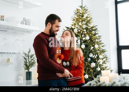 Ein verliebtes Paar umarmt und küsst sich in der Nähe des Weihnachtsbaums im dekorierten Inneren des Weihnachtsbaums zu Hause an einem Weihnachtsurlaub im Winter. Ein Mann Stockfoto