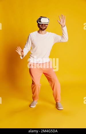 Studioaufnahmen eines jungen weißen Mannes, der lässige Kleidung trägt und eine VR-Brille verwendet, isoliert auf gelbem Hintergrund. Emotionen, Spiele, Technik, Werbung Stockfoto