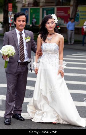 Vietnamesisches Paar mit Blumenstrauß, das für ein Hochzeitsfoto posiert und die Straße überquert, im Zentrum von Ho Chi Minh City, Vietnam Stockfoto