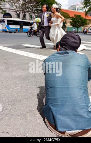 Ein vietnamesischer Hochzeitsfotograf fotografiert ein Paar, das auf der anderen Straßenseite im Zentrum von Ho-Chi-Minh-Stadt, Vietnam, läuft Stockfoto