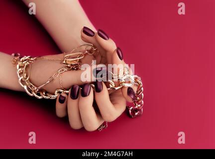 Weibliche Hände mit trendigen dunklen Nagelmustern in goldenen Armbändern auf trendy magentafarbener Farbe im Hintergrund des Jahres 2023. Luxuriöses Manikürkonzept. Stockfoto