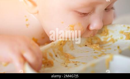 Das kleine Mädchen nimmt Essensreste vom weißen Teller und isst mit Begeisterung und besorgtem Ausdruck. Das Kind schmeckt Essen an den Händen und bekommt Gesicht und Finger di Stockfoto