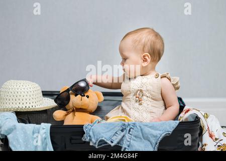 Ein glückliches Mädchen, das im Reisekoffer sitzt, trägt eine Sonnenbrille auf einem Bärenspielzeug. Ein Kind mit amüsiertem Gesichtsausdruck bereitet sich gern auf den Sommerurlaub vor, Selekti Stockfoto