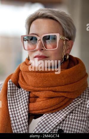 Portrait Woman kurze blonde Haare in warmem, ockerfarbenen Strickschal, zweireihiger Jacke und modischer Brille. Herbst oder Frühling im Modestil, weiblich Stockfoto