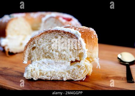 Roscon de reyes gefüllt mit Sahne, Brötchen oder Kuchen, typisch für Spanien, am 3. Königstag mit einer Portion auf schwarzem Teller und schwarzem Hintergrund. Stockfoto