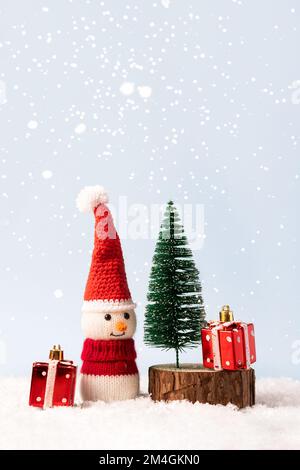 Weihnachtsmärchen-Komposition eines gestrickten Schneemanns mit Weihnachtsmann-Hut und -Pullover. Kleiner Weihnachtsbaum mit zwei roten Geschenken Stockfoto