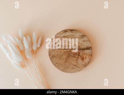 Holzbraunes rundes Podium und Strauß aus trockenem Lagurus auf beigefarbenem Hintergrund. Ort, Hintergrund für Kosmetik. Draufsicht. Speicherplatz kopieren Stockfoto