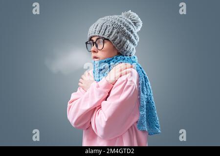 Krankheit und Grippe. Porträt einer jungen Frau mit Brille, Hut und Schal wärmt sich und atmet. Grauer Hintergrund. Das Konzept von Energy cr Stockfoto