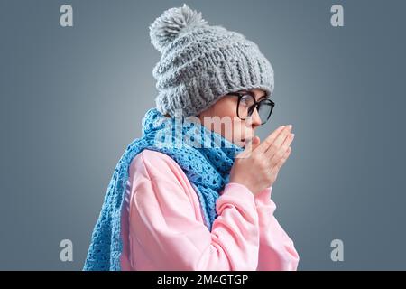 Porträt einer jungen kaukasischen Frau mit Brille, Hut und Strickschal wärmt sich. Grauer Hintergrund. Das Konzept der Grippe und Wintersaison. Stockfoto