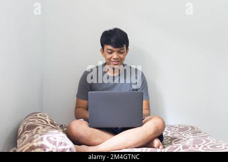 Porträt ein asiatischer Mann lächelt und schaut auf einen Laptop in einem Schlafzimmer für die Arbeit von zu Hause Konzept mit natürlichem Licht Stockfoto