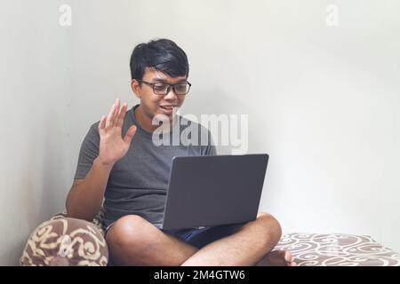 Porträt asiatische Brille Mann hebt seine Hand und schaut auf Laptop in einem Schlafzimmer für Arbeit von zu Hause Konzept mit natürlichem Licht isolierten weißen Backgro Stockfoto