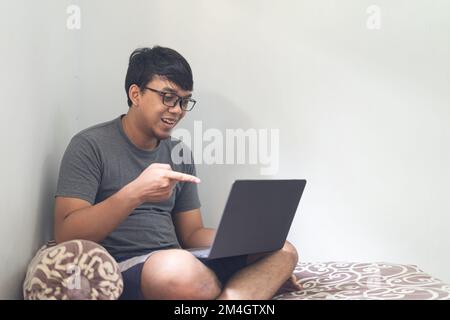Porträt asiatische Brille der Mann zeigt und schaut auf einen Laptop in einem Schlafzimmer für die Arbeit von zu Hause Konzept mit natürlichem Licht Stockfoto