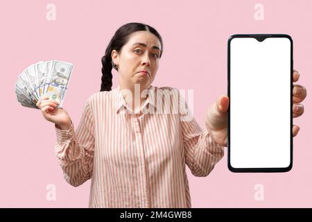 Banking-App. Eine zweifelhafte junge weiße Frau zeigt ein Smartphone mit einem weißen Bildschirmmodell und hält einen Geldfan auf pinkfarbenem Hintergrund. Das Konzept Stockfoto