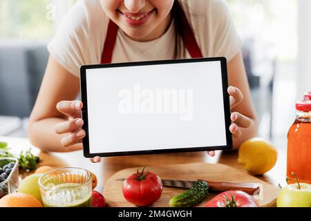 Frau Hände halten Tablet, zeigt weiße leere Bildschirmmodelle in der Küche, während gesundes Gemüse-Frühstück kocht. Online-Lebensmitteleinkäufe. Werbung mit App, Kochbuch, Rezepte für Diätpläne Stockfoto