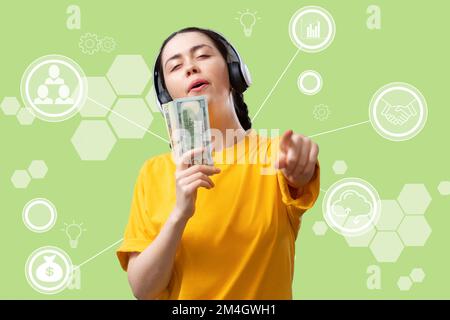 Eine Lotterie gewinnen. Eine junge, weiße, glückliche Frau mit Kopfhörern, die ein Bündel Dollar als Mikrofon singt. Grüner Hintergrund mit digitalen Infografiken. T Stockfoto