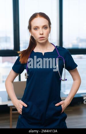 Vertikales Porträt einer selbstbewussten hübschen Ärztin, die medizinische Uniform trägt und auf die Kamera schaut, die auf dem Hintergrund des Fensters steht. Stockfoto